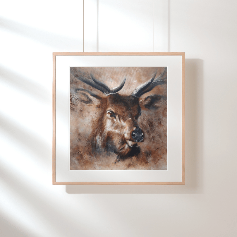 A framed art print of a deer head with antlers, shown hanging on a white wall with soft diagonal shadows. The print features a textured brown and beige painting with a white mat and a light natural wood frame.