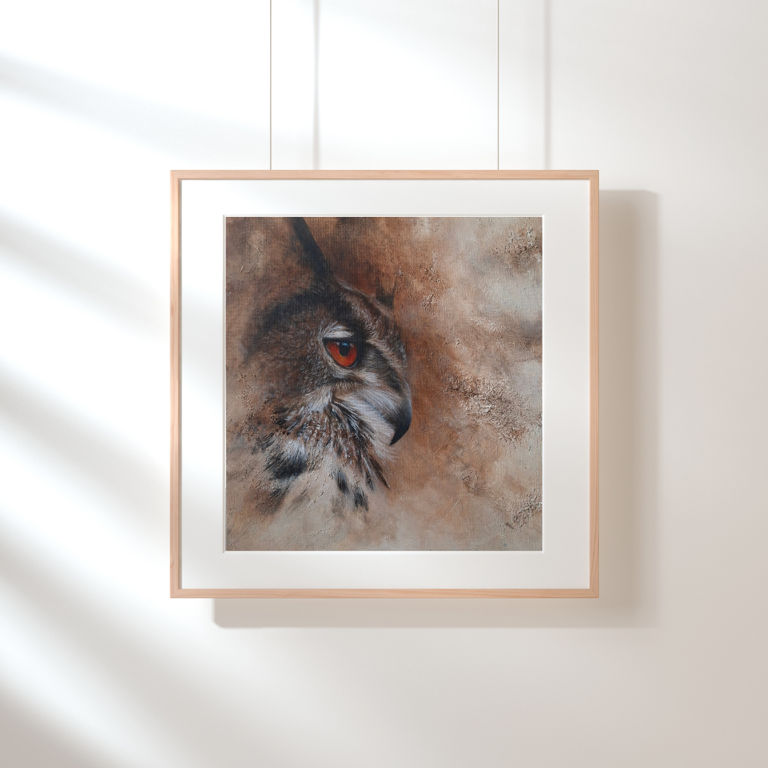 A framed art print of an owl's face, featuring a realistic brown and gray feathered texture with a striking red eye, presented in a light wood frame with a white mat against a white wall.