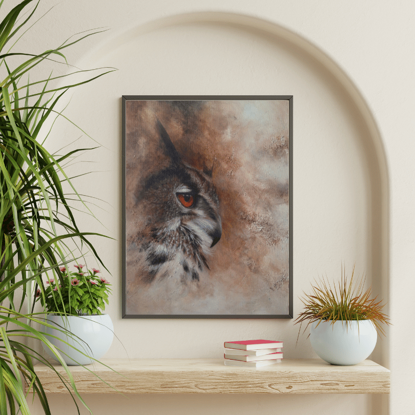 A framed art print of an owl's face with a warm orange eye, displayed on a light wood shelf in a styled room setting with potted plants.
