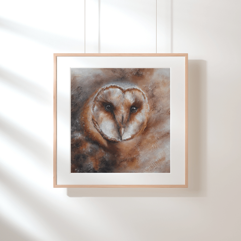 A framed art print of a barn owl hanging on a white wall with soft window shadows. The print features a textured, painterly depiction of the owl in warm brown, beige, and white tones, enclosed in a light wood frame with a white mat.